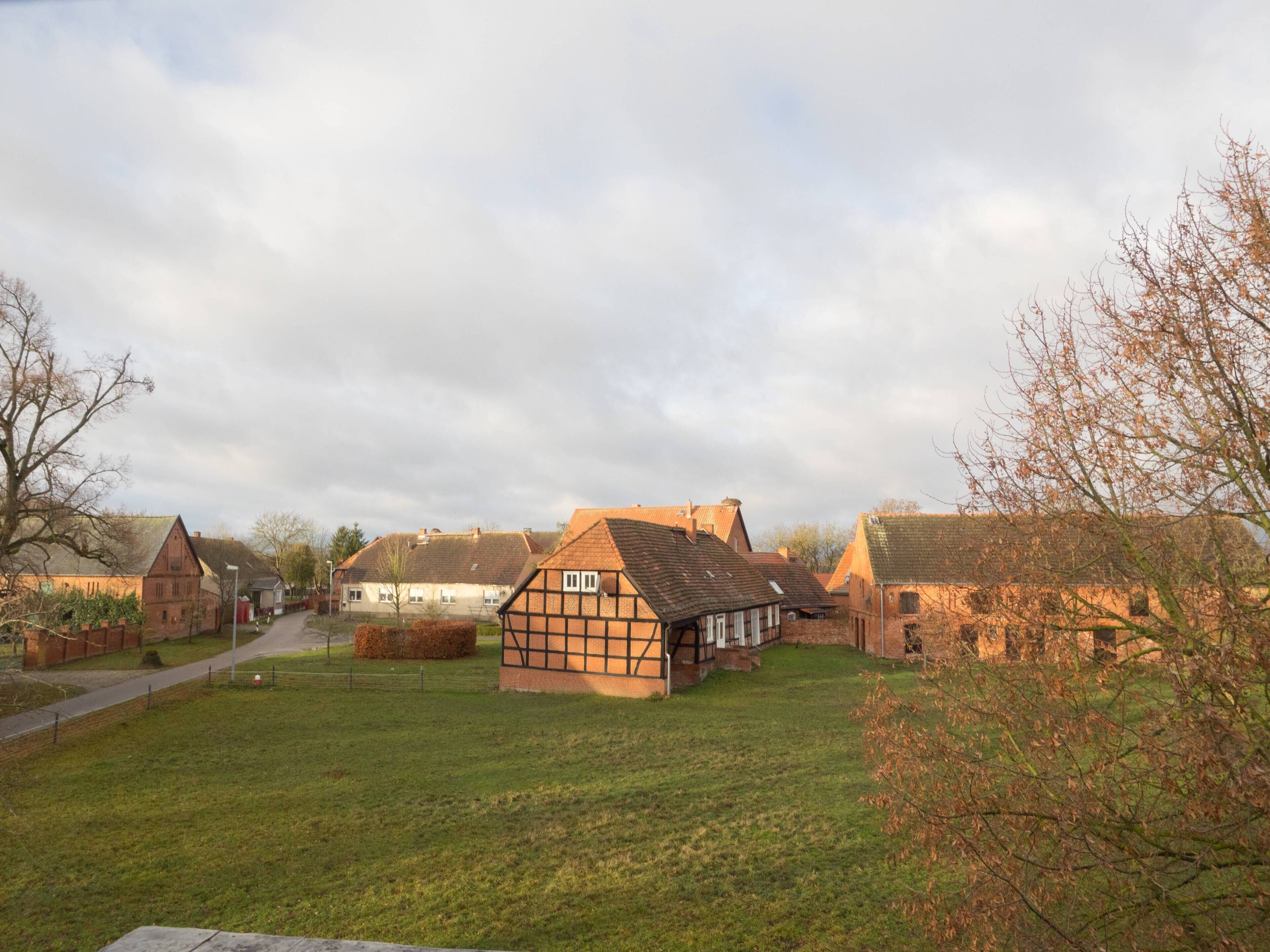 Une maison traditionnelle à colombages, avec un toit de chaume, entourée d’une pelouse et d’autres maisons similaires.