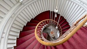 Una majestuosa escalera en espiral con barandilla de madera y peldaños rojos, que conduce a un rellano con techo decorativo y una ventana.