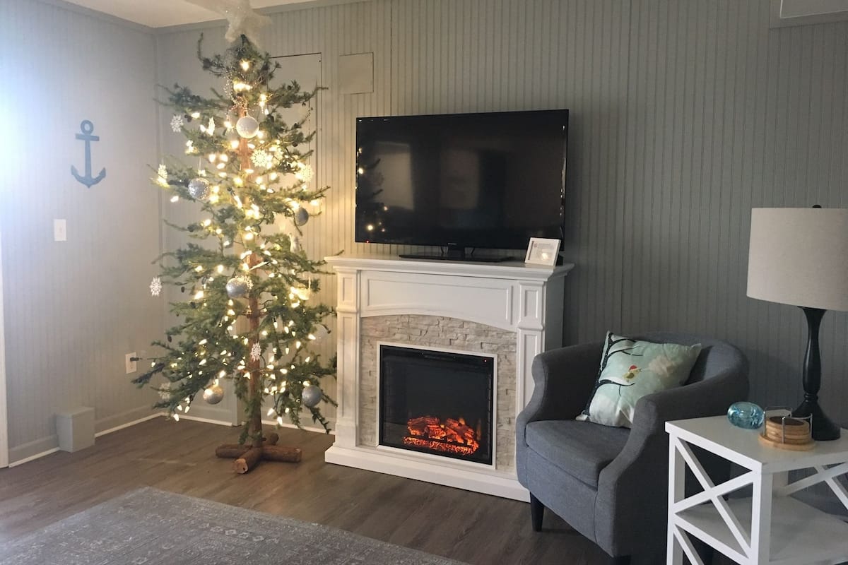 A living room with a fireplace, a television mounted above it, a Christmas tree, and a grey armchair.