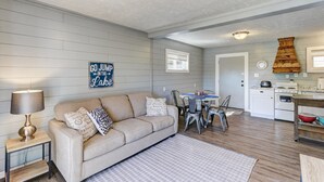 A living room with a sofa, a coffee table, and a dining area with chairs and a table. There is a kitchen area with a stove and cabinets. - Cottages at Fair Haven Cove (Cape Fair)
