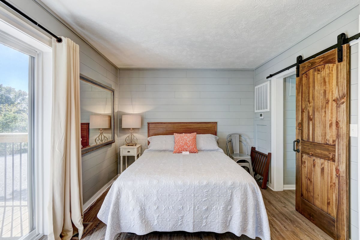 A bedroom with a large bed, a wooden door, a nightstand, a lamp, and a view of the outdoors.