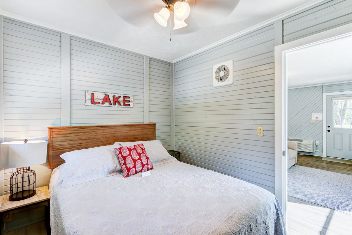 A bedroom with a bed, a nightstand, a wall-mounted fan, and a door leading to another room.