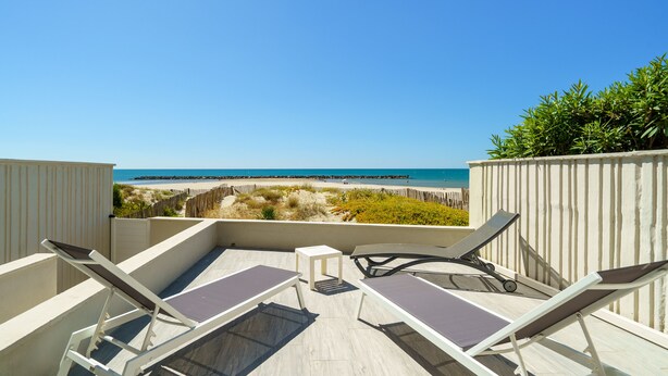 Terraza en la azotea con dos tumbonas y una pequeña mesa, con vistas a una playa con valla de madera y vegetación.