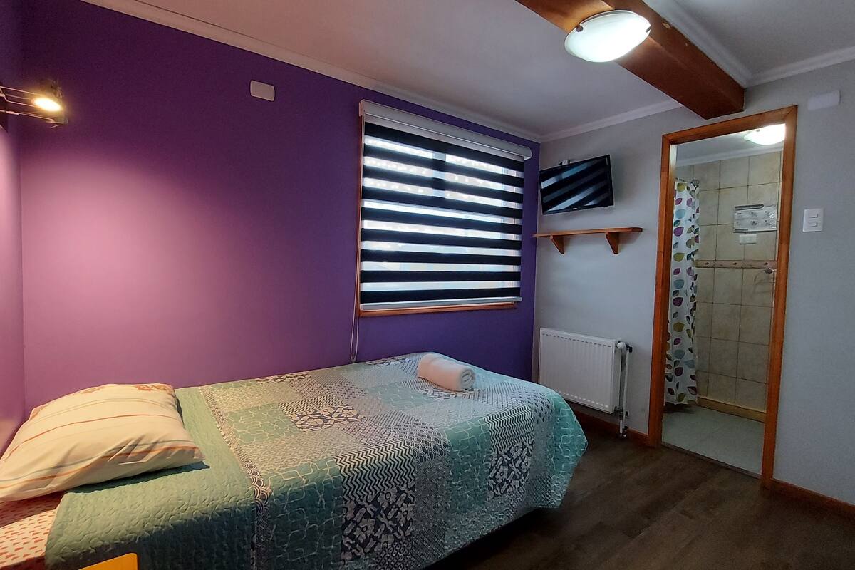 A bedroom with a single bed, purple walls, a window with blinds, a mounted TV, and a wooden ceiling beam.