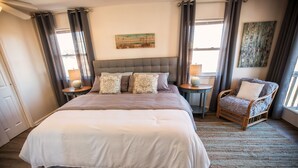Quarto com uma cama grande, duas mesas de cabeceira com abajures, uma cadeira e obras de arte nas paredes.
