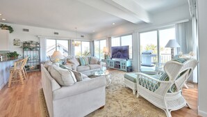 A spacious living room with a large sofa, a white wicker chair, and a television. There is a kitchen area with a dining table and chairs, and a balcony with a grill. - 2027_Ocean View Completely upgraded-New everything_ 2027 Rates (Harvey Cedars)