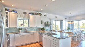 A modern kitchen with white cabinets, a granite countertop, and a central island with a sink. There are potted plants and decorative items on the shelves. - 2027_Ocean View Completely upgraded-New everything_ 2027 Rates (Harvey Cedars)