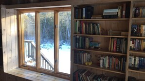 Een kamer met een groot raam dat uitkijkt op een besneeuwde veranda, houten boekenkasten vol boeken en een rustiek houten interieur.
