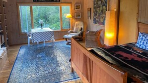 Een gezellige kamer met een houten bed, een stoel, een tafel met een lamp en een groot raam met uitzicht op groen.