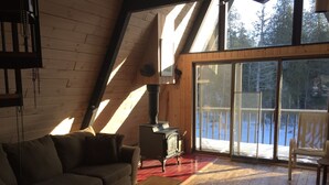 Een gezellig cabin interieur met een houten vloer, een bank, een tafel, een open haard en een grote schuifdeur die uitkomt op een terras met uitzicht op sneeuw.