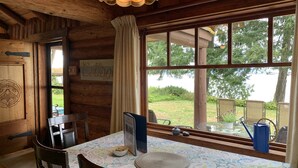 Interior de uma cabana de madeira com uma mesa de jantar, cadeiras, uma janela com vista para vegetação e um letreiro com a palavra 'Cottage'.