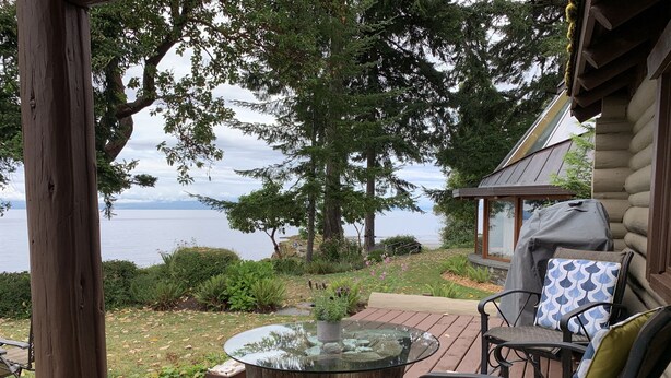 Um deck de madeira com mesa e cadeiras de tampo de vidro, com vista para um lago e uma cabana.