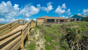 A wooden boardwalk leading to a building with a sandy beach and grassy dunes in the background.