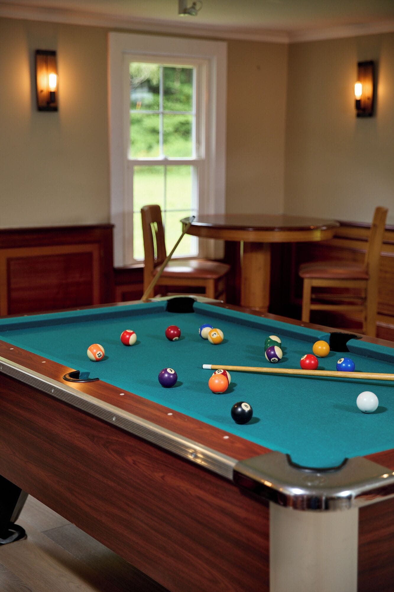 A pool table with balls and a cue stick, a wooden chair, and a table with chairs in the background.