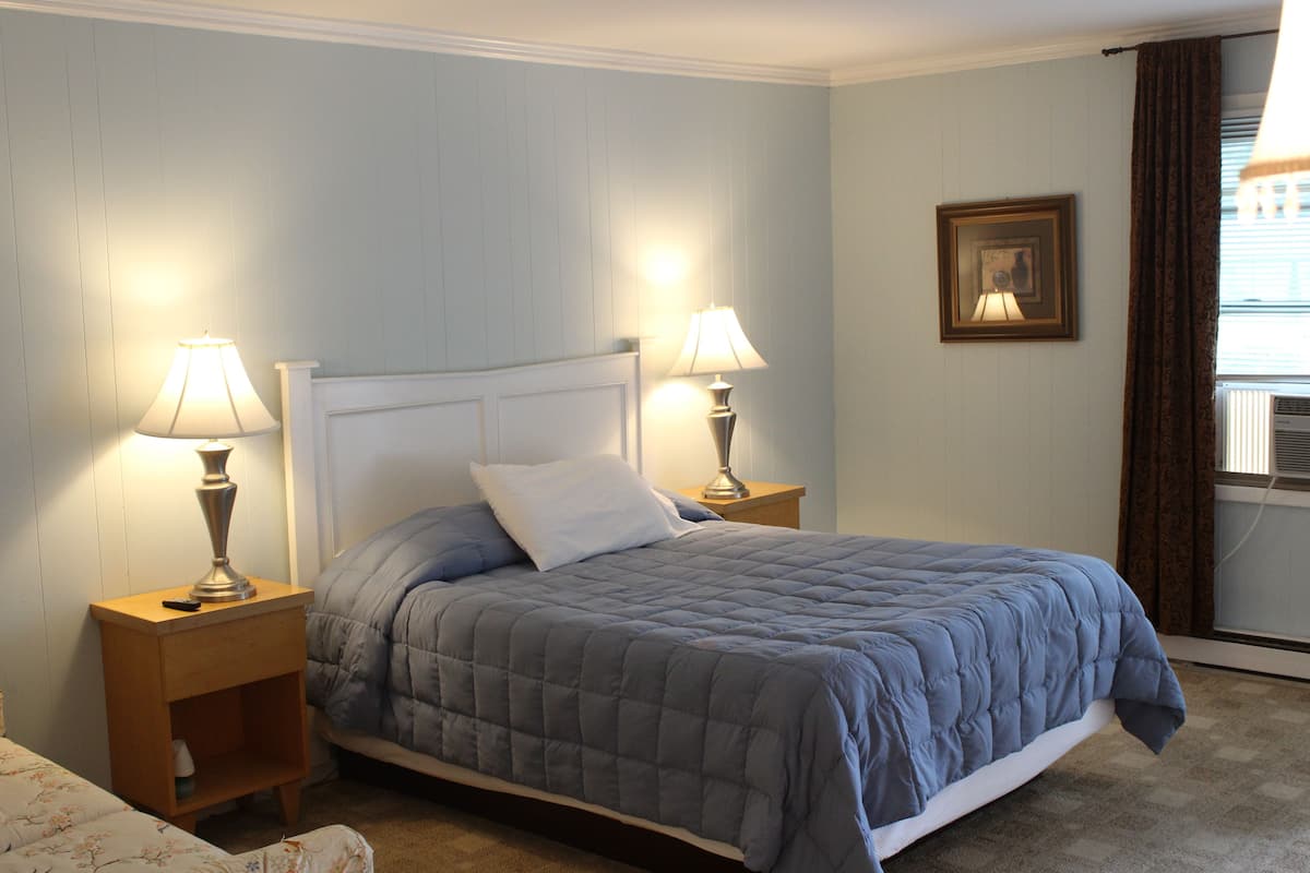 A bedroom with a bed, two bedside lamps, a nightstand, a framed picture on the wall, and a window with curtains.