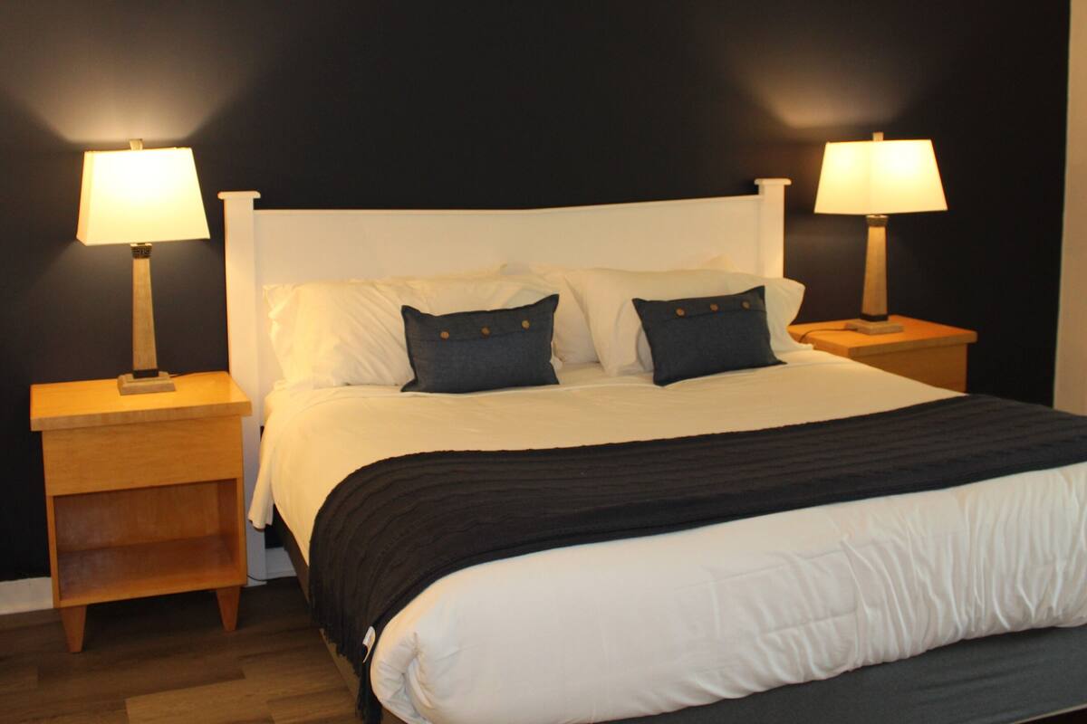 A neatly made bed with white bedding and a black throw blanket, flanked by two bedside tables with lamps, against a dark wall.
