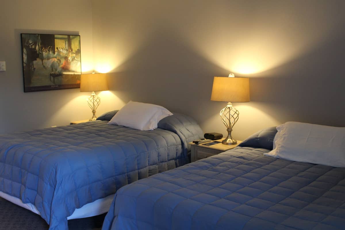A hotel room with two beds, each with a blue quilted bedspread and white pillows. There are two bedside lamps with lampshades, a painting on the wall, and a small table between the beds.