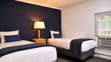 A hotel room with two beds, a nightstand with a lamp, a window with blinds, and a radiator.