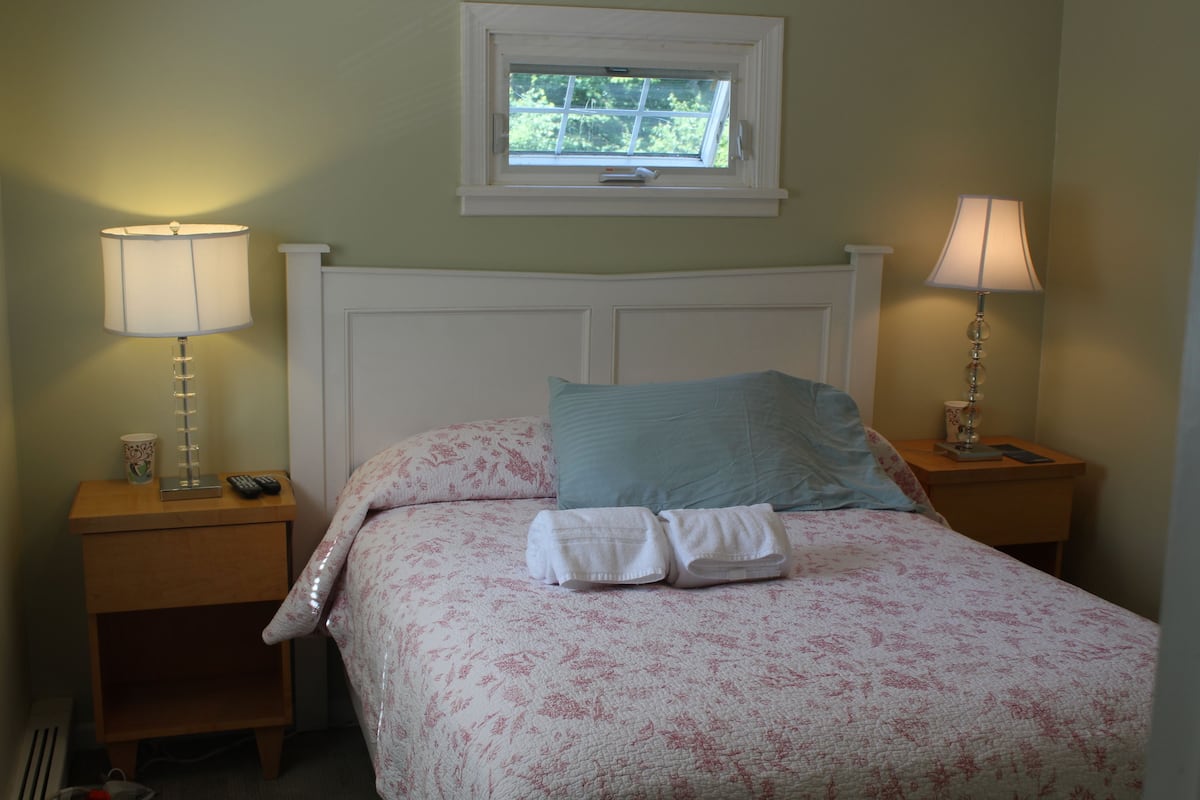 A neatly made bed with a floral bedspread, two pillows, and folded towels on a wooden nightstand.