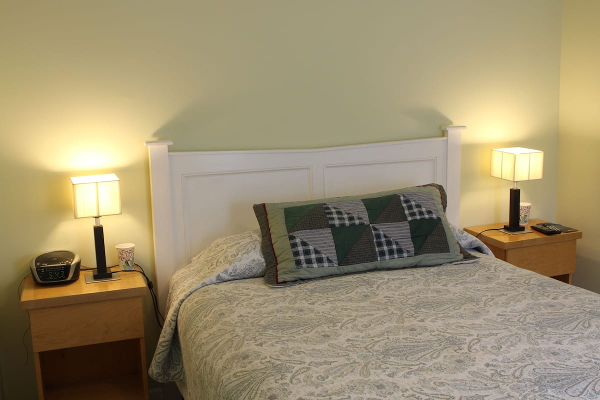 A neatly made bed with a patterned bedspread, two bedside lamps, a clock, and a cup on the nightstand.