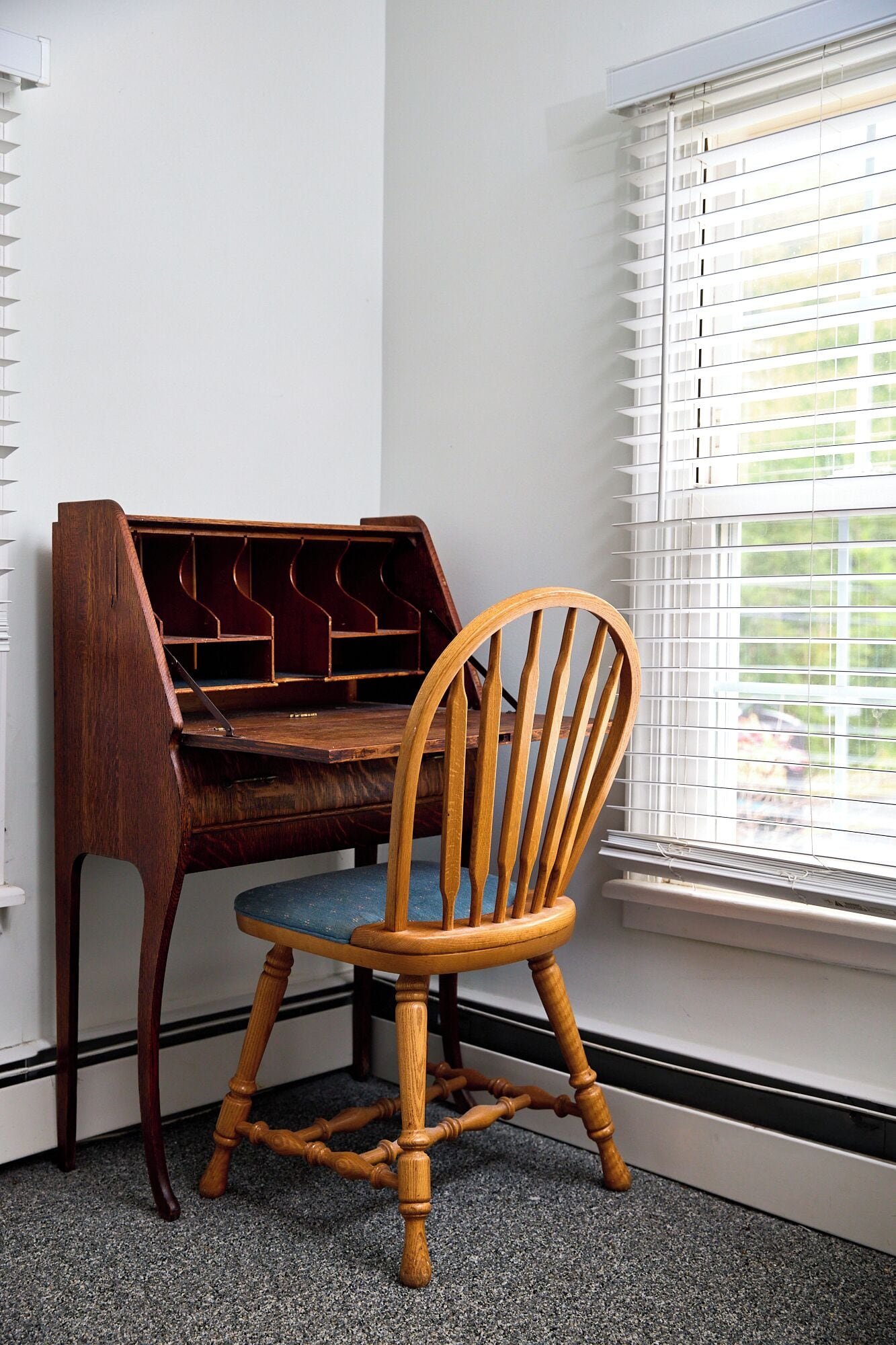 Une chaise de bureau en bois, dotée d’un siège et d’un dossier rembourrés, placée devant une fenêtre munie de stores.
