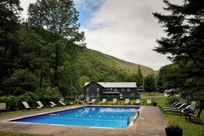 A swimming pool with lounge chairs, a large building, and a forested hill in the background.