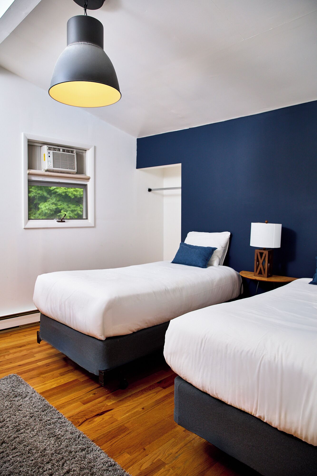A bedroom with two beds, a window with an air conditioner, a lamp on a side table, and a ceiling light.