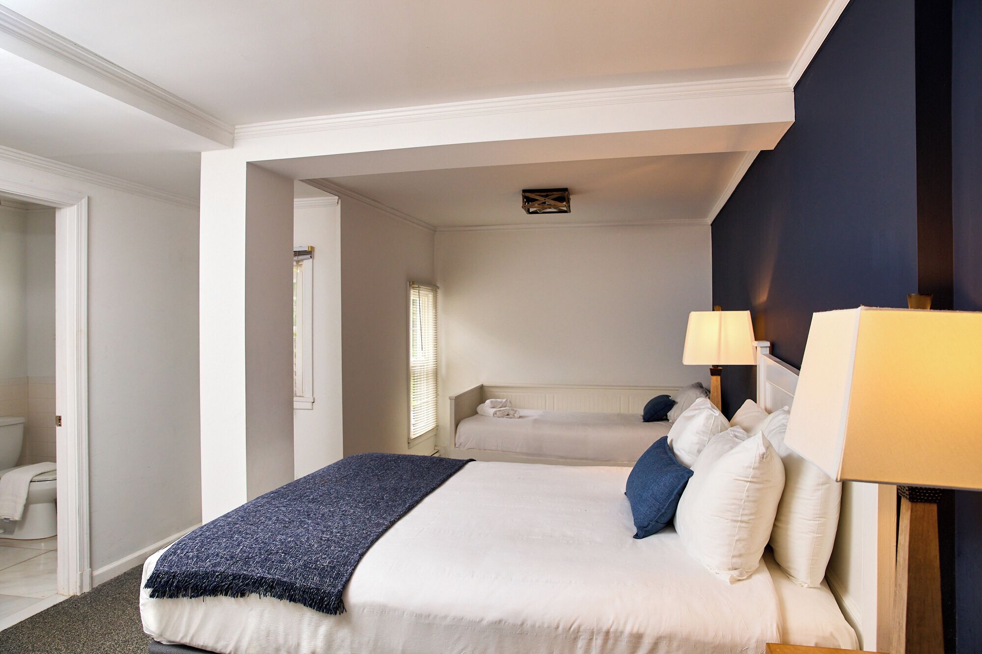 A hotel room with a bed, bedside lamps, a window, and a view of another room.