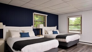A hotel room with two beds, a nightstand, a lamp, and a window with blinds.