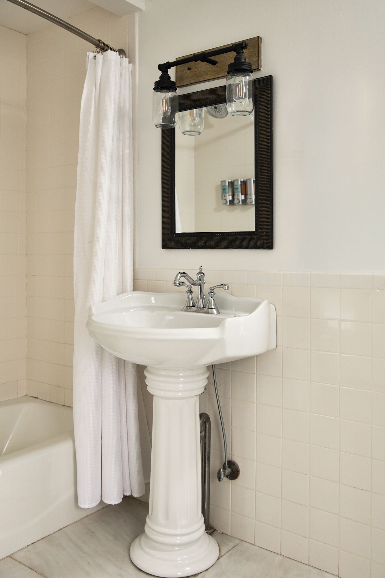 Une salle de bain avec un lavabo sur colonne, un rideau de douche, un miroir avec un cadre en bois et une étagère contenant des articles de toilette.