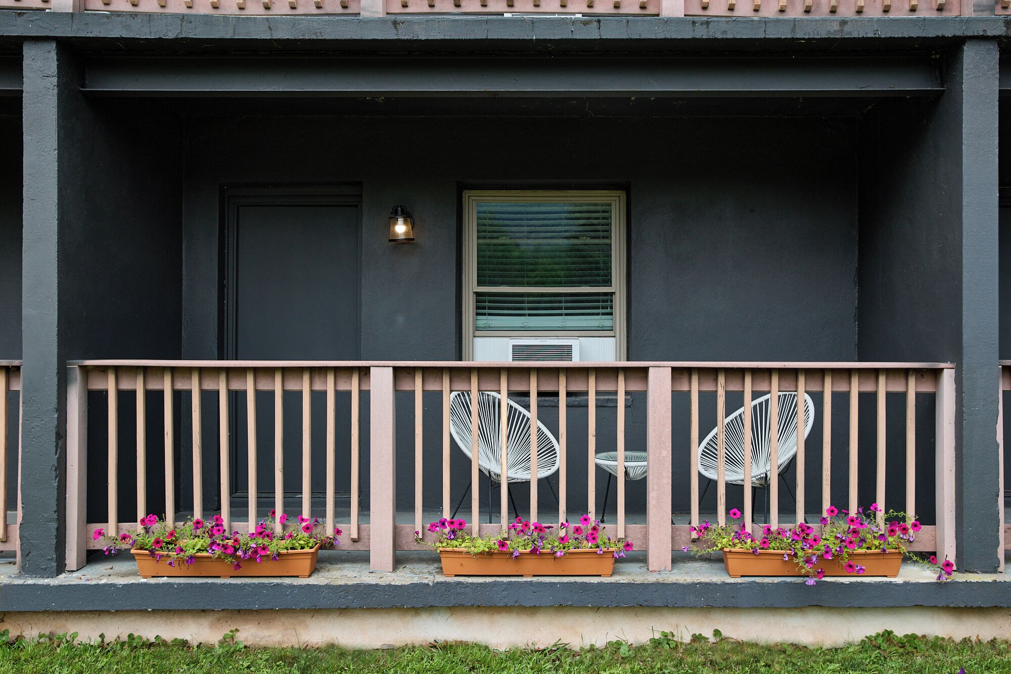 A building facade with a dark exterior, a window with blinds, a small porch with a railing, and flower planters on the porch.