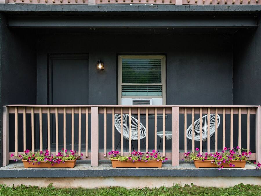En byggnadsfasad med en mörk exteriör, ett fönster med persienner, en liten veranda med ett räcke och blomlådor på verandan.