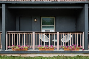 A building facade with a dark exterior, a window with blinds, a small porch with a railing, and flower planters on the porch.