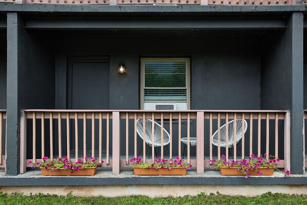 A building facade with a dark exterior, a window with blinds, a small porch with a railing, and flower planters on the porch.