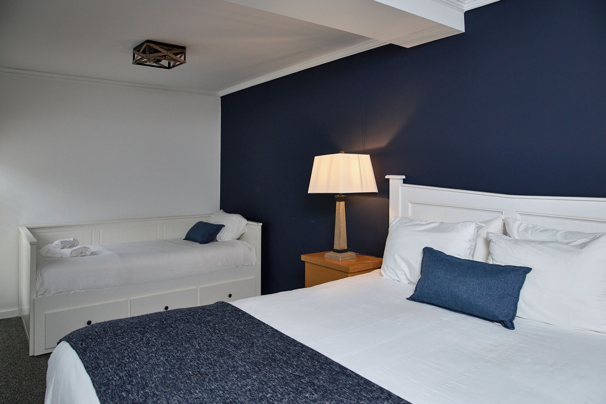 Habitación de hotel con cama blanca, manta azul, mesita de noche con lámpara y pared azul oscuro de acento.