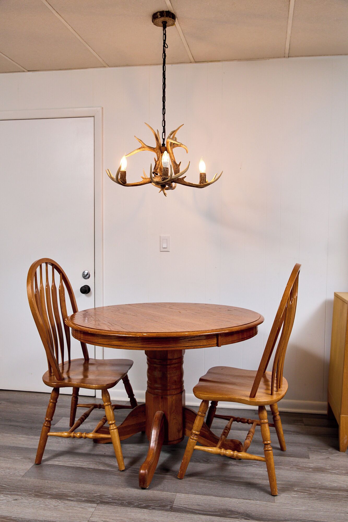 Un comedor con una mesa redonda de madera y cuatro sillas, una araña con luces, y una puerta blanca al fondo.
