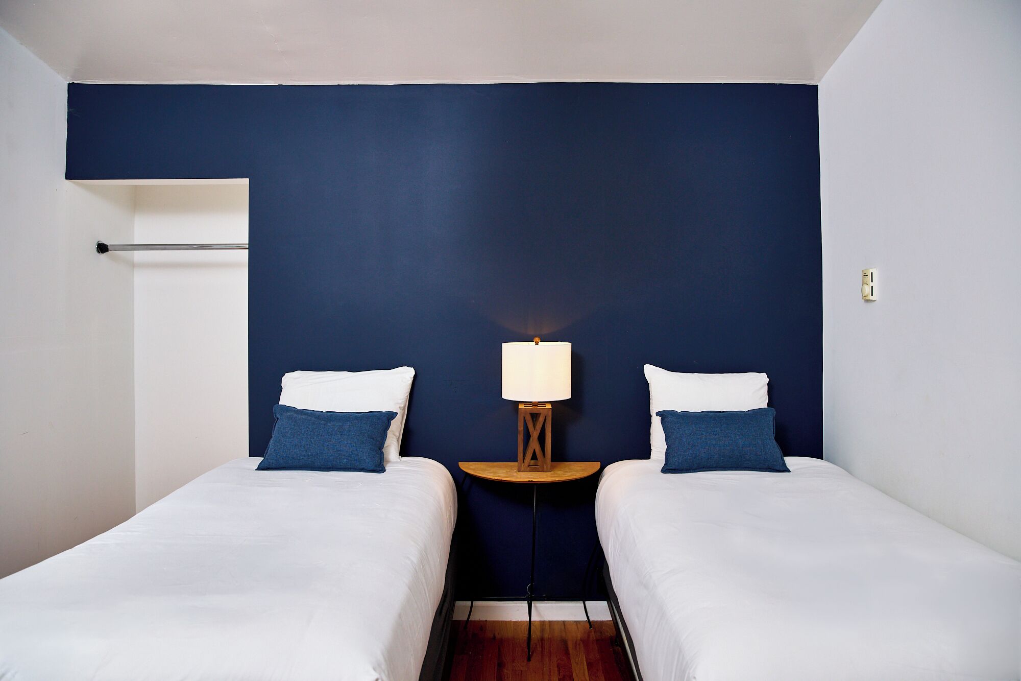 A hotel room with two single beds, a nightstand with a lamp, and a dark blue accent wall.