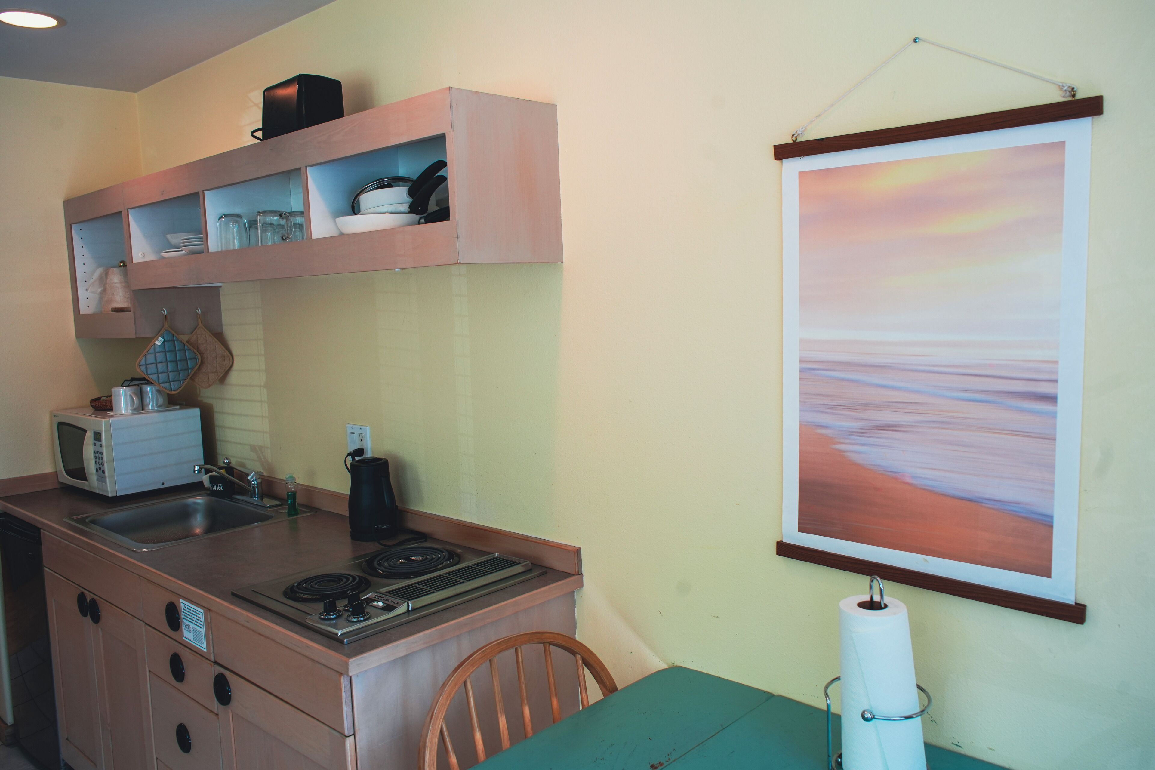 A compact kitchen with a microwave, stove, sink, and a painting of a sunset on the wall.