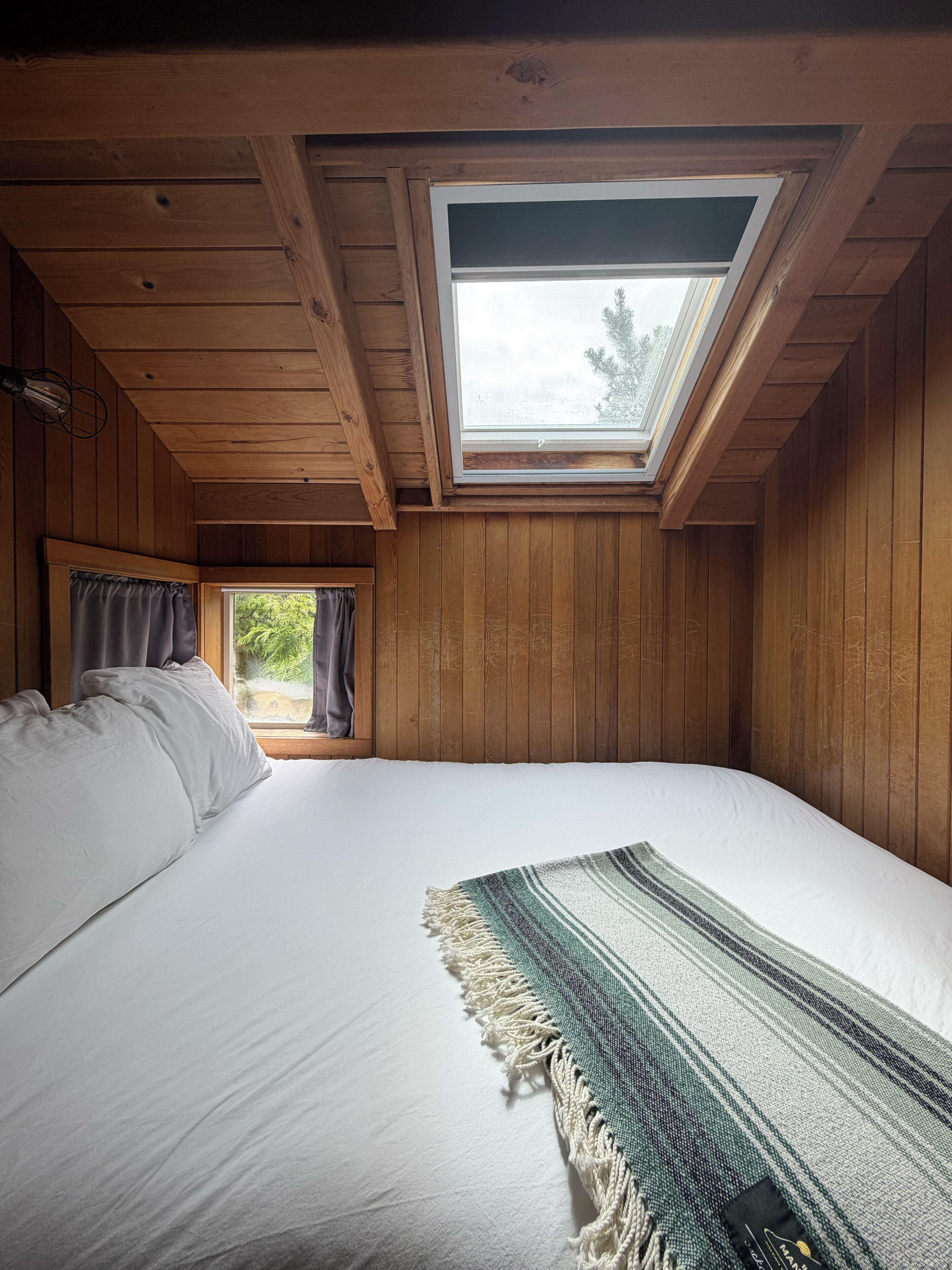 Una habitación con tragaluces, paredes de madera, una cama con sábanas blancas y una manta a rayas.