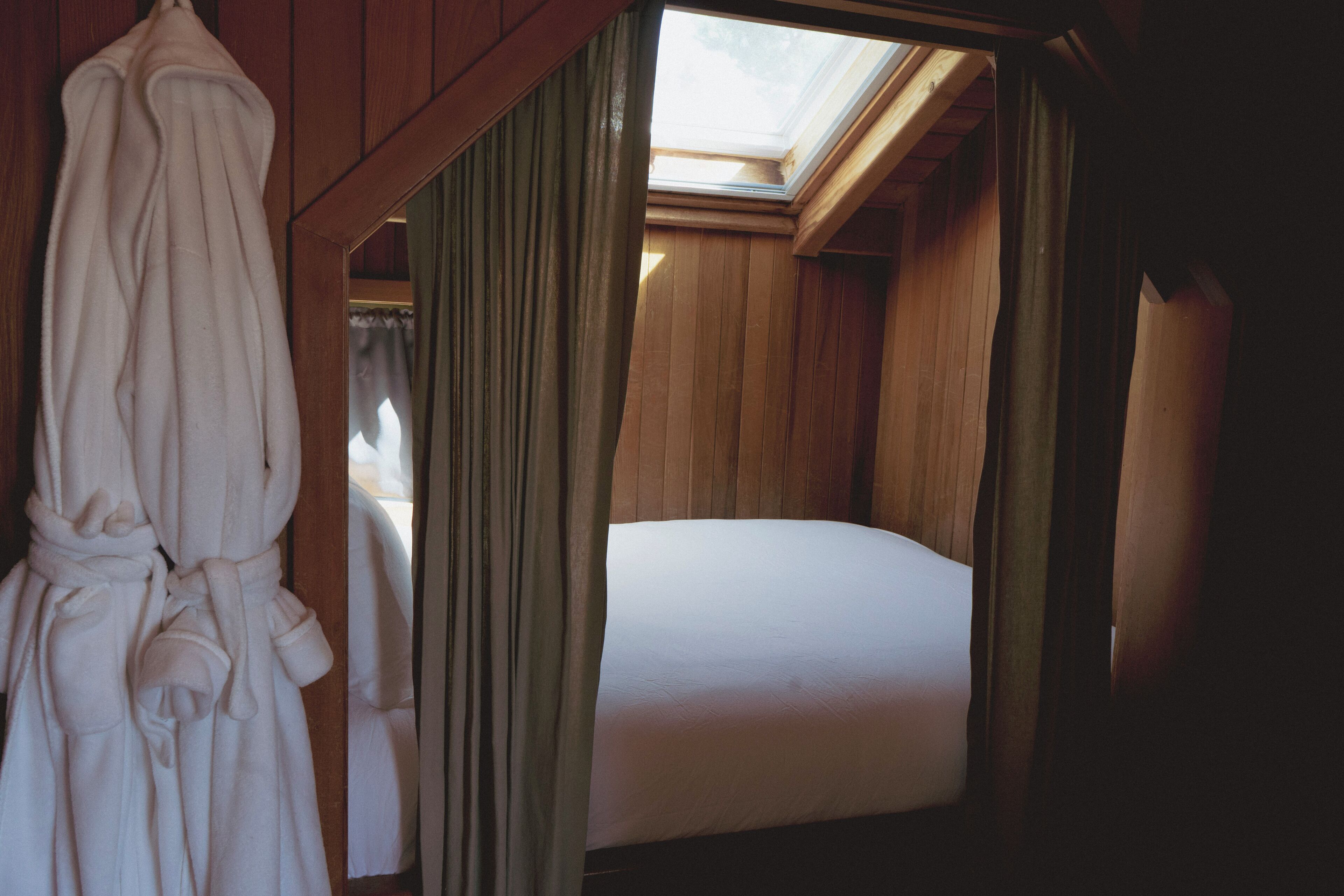 Une pièce en bois avec un lit, une robe suspendue au mur et une lucarne.