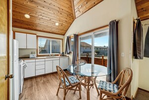 Cocina compacta con gabinetes blancos, una mesa de comedor de vidrio, sillas de mimbre y un ventanal con cortinas.
