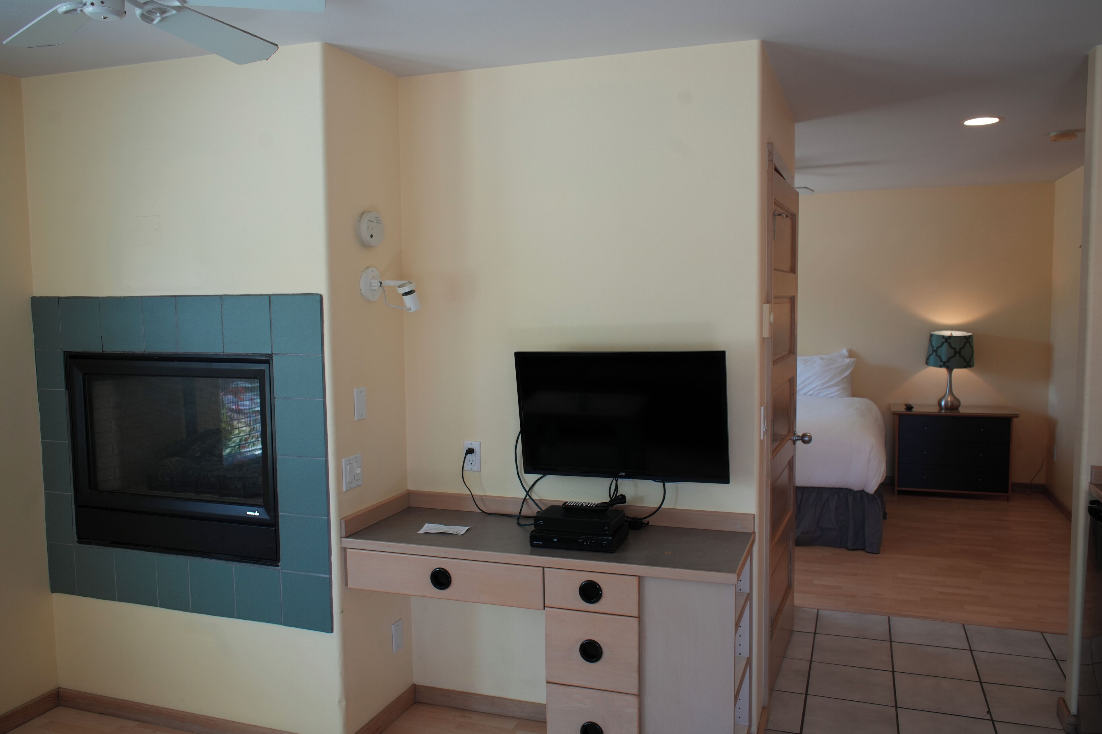 Quarto compacto com televisão numa escrivaninha, uma janela pequena e uma cama ao fundo.