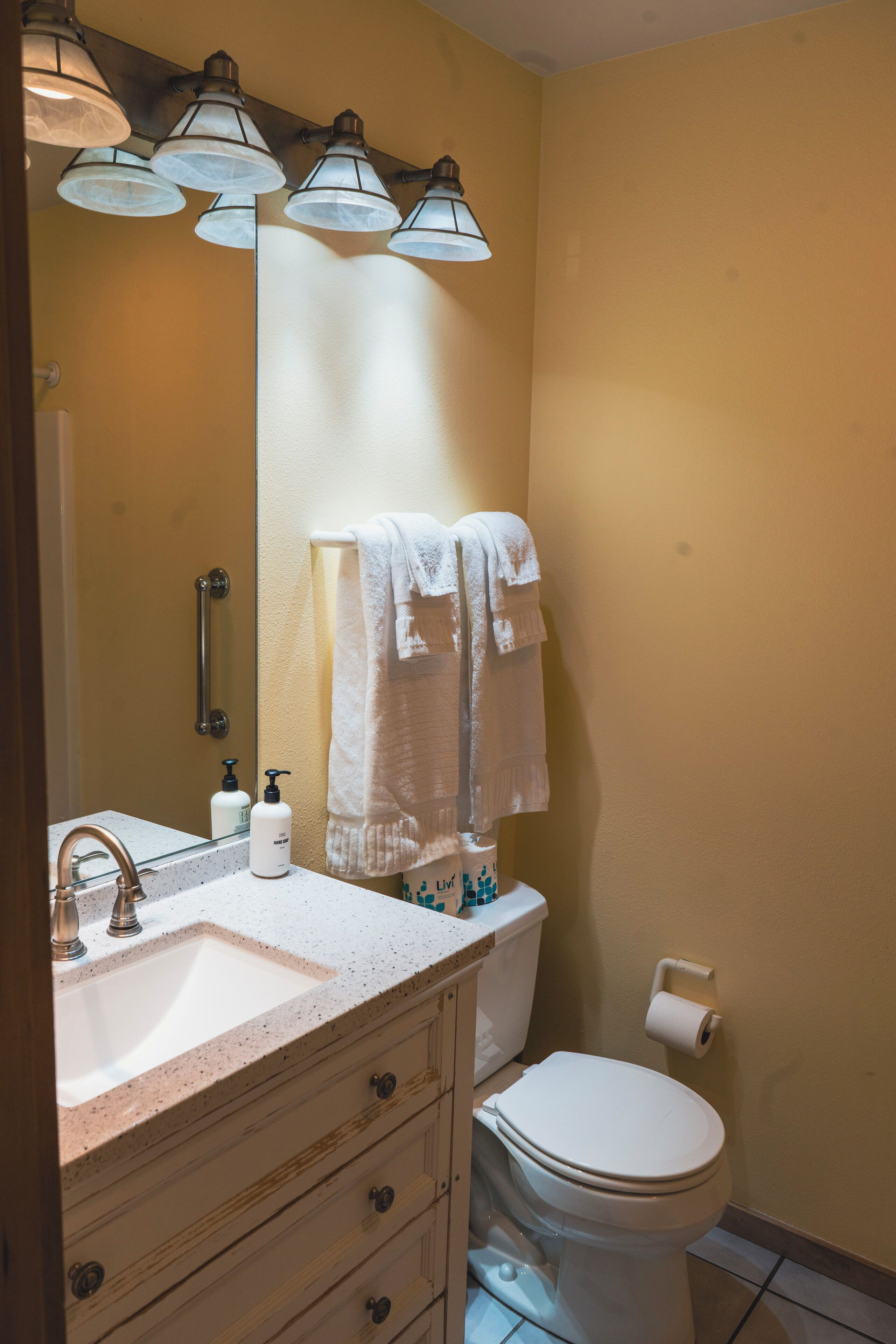 A bathroom with a sink, mirror, towel rack, toilet, and a roll of toilet paper.