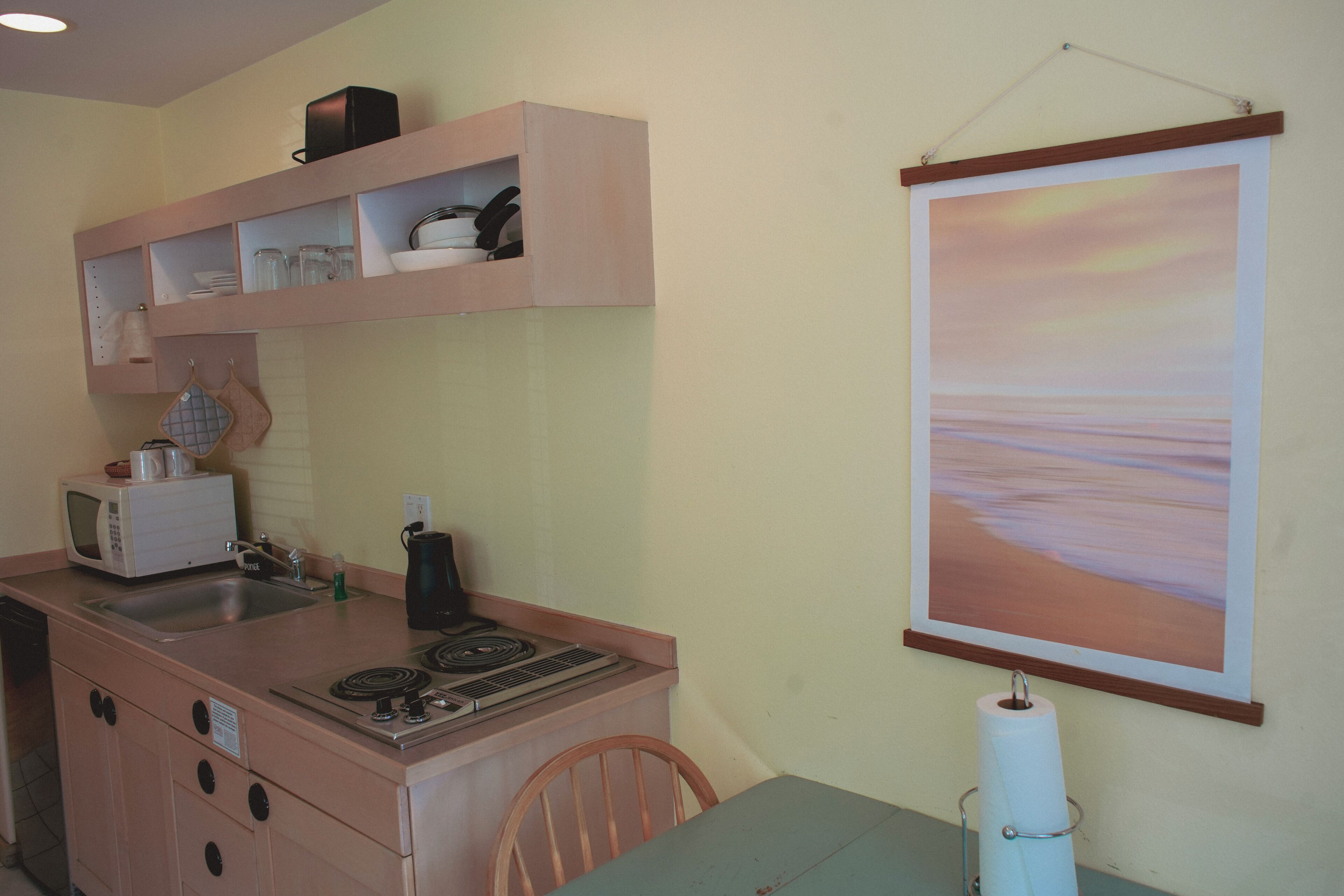 A compact kitchen with a microwave, sink, and stove, a wooden chair, and a framed painting on the wall.