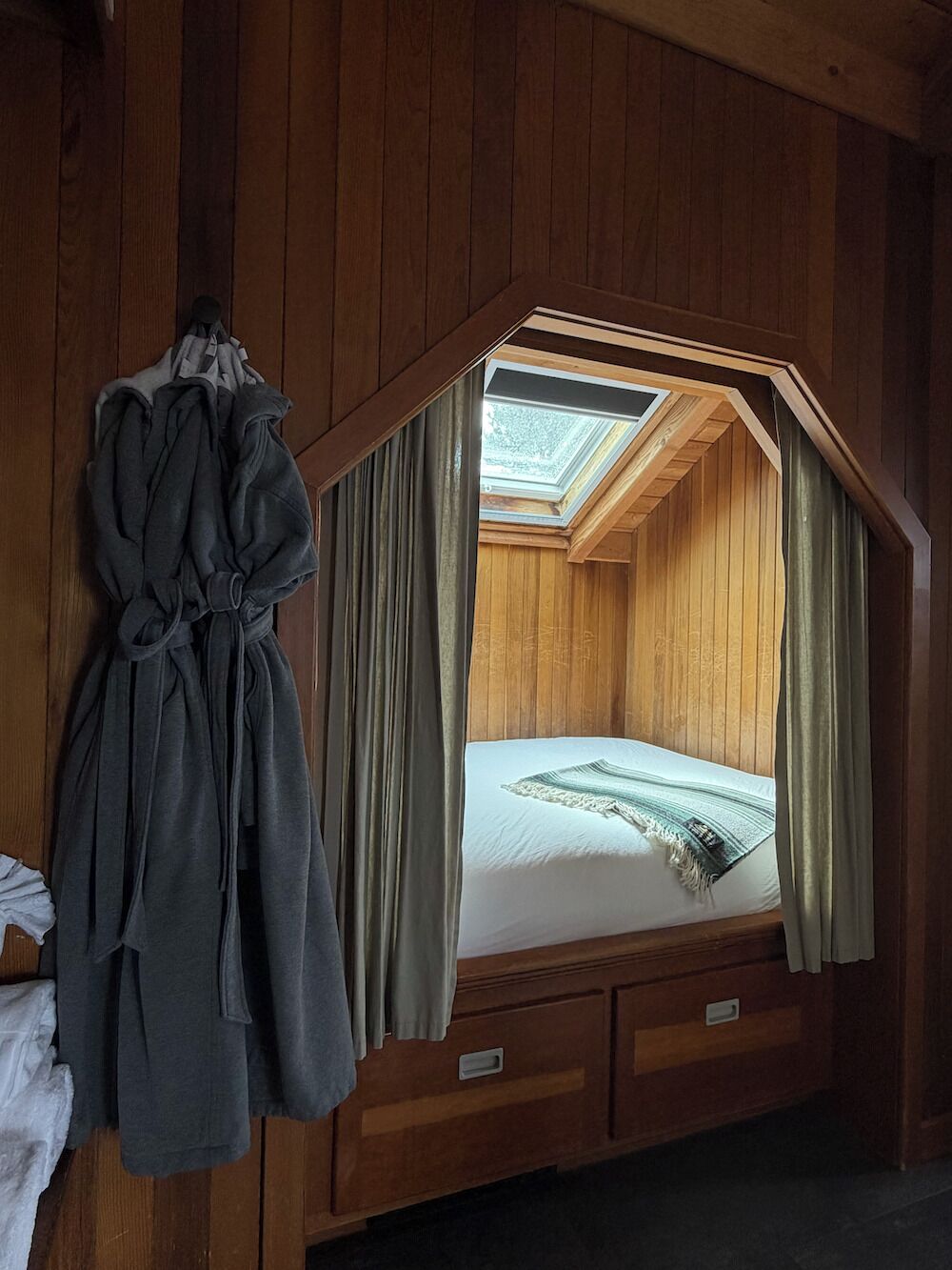 A wooden room with a bed, a robe hanging, and a skylight.