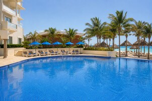A clear blue pool with lounge chairs and umbrellas, surrounded by palm trees and a building with balconies.