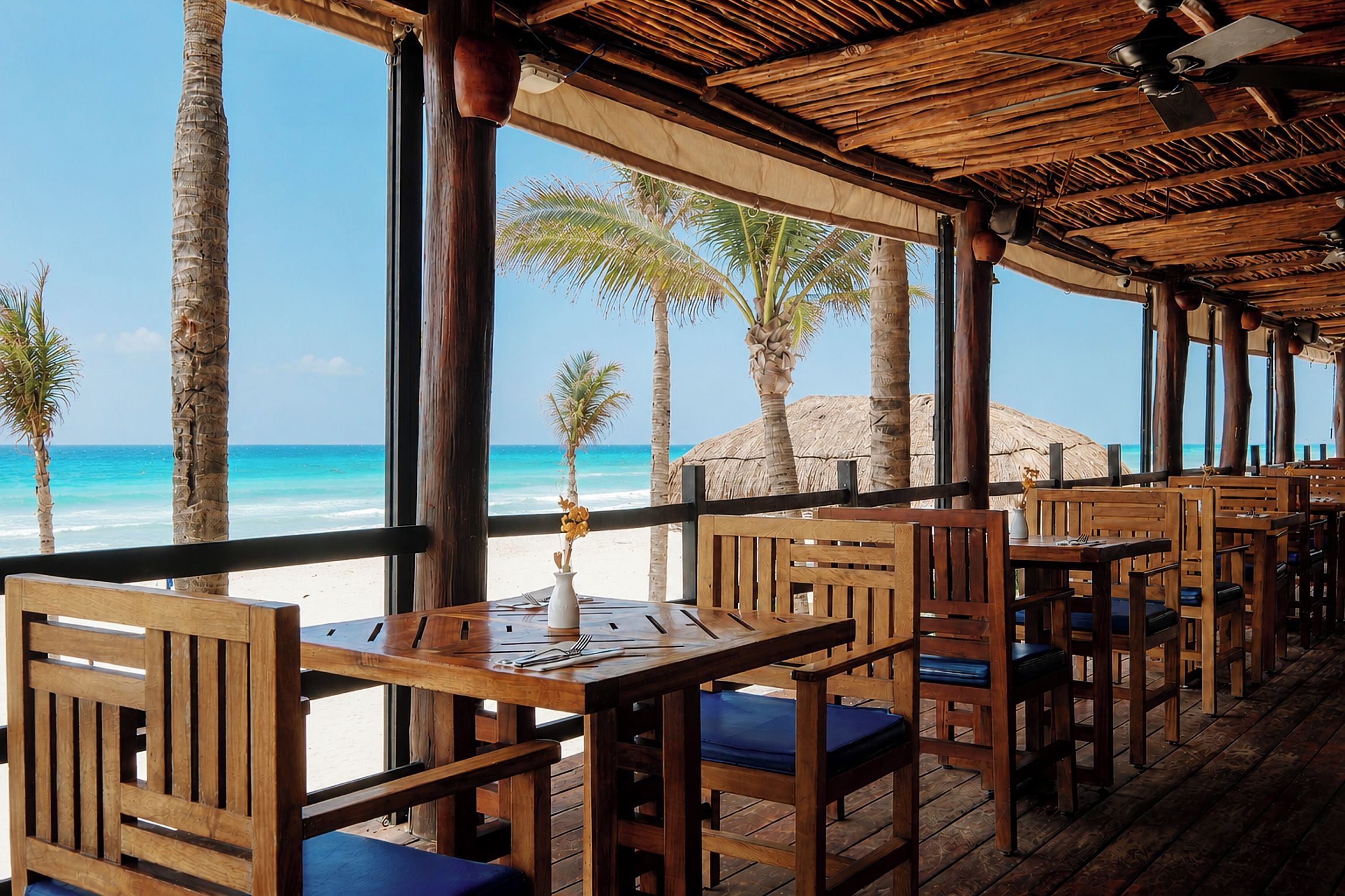 Un restaurante al aire libre con mesas y sillas de madera, con vistas a una playa con palmeras y el océano.