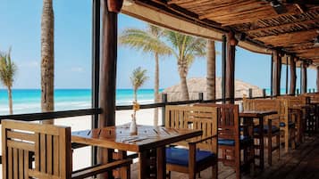 Un restaurante al aire libre con mesas y sillas de madera, con vistas a una playa con palmeras y el océano.