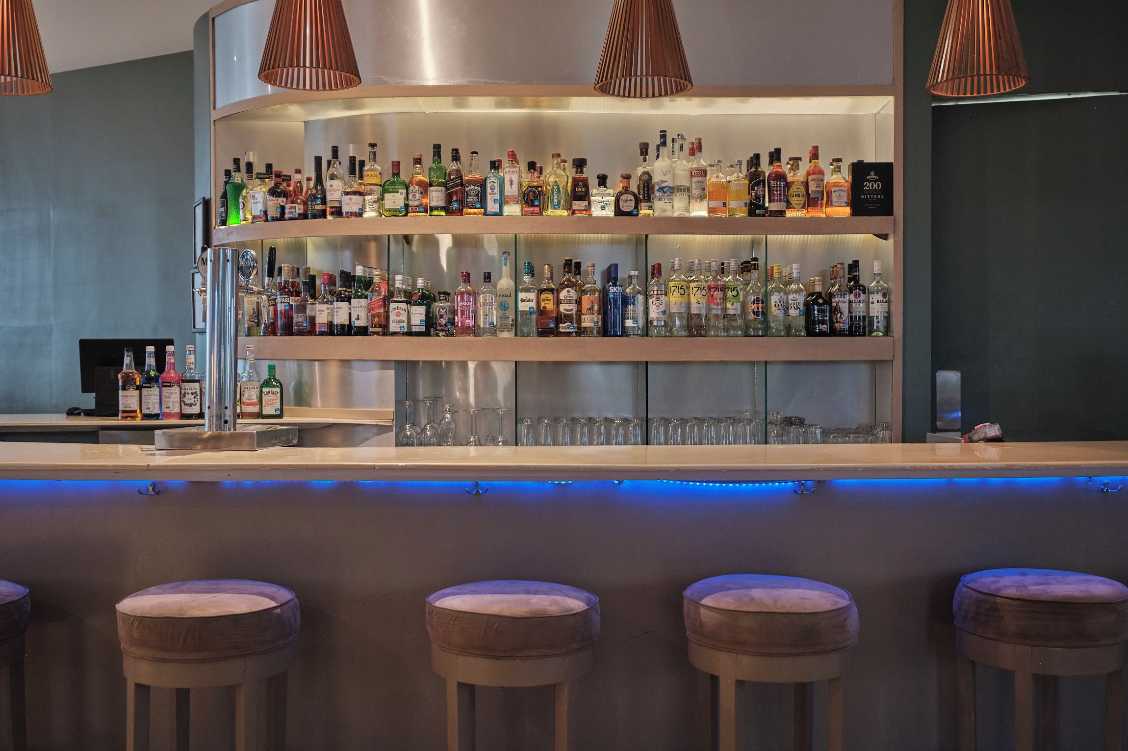 A bar area with multiple shelves of liquor bottles, a bar counter, and three cushioned stools.