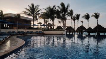 Zona de piscina con sillas de descanso y sombrillas, rodeada de palmeras y un edificio de diseño moderno.